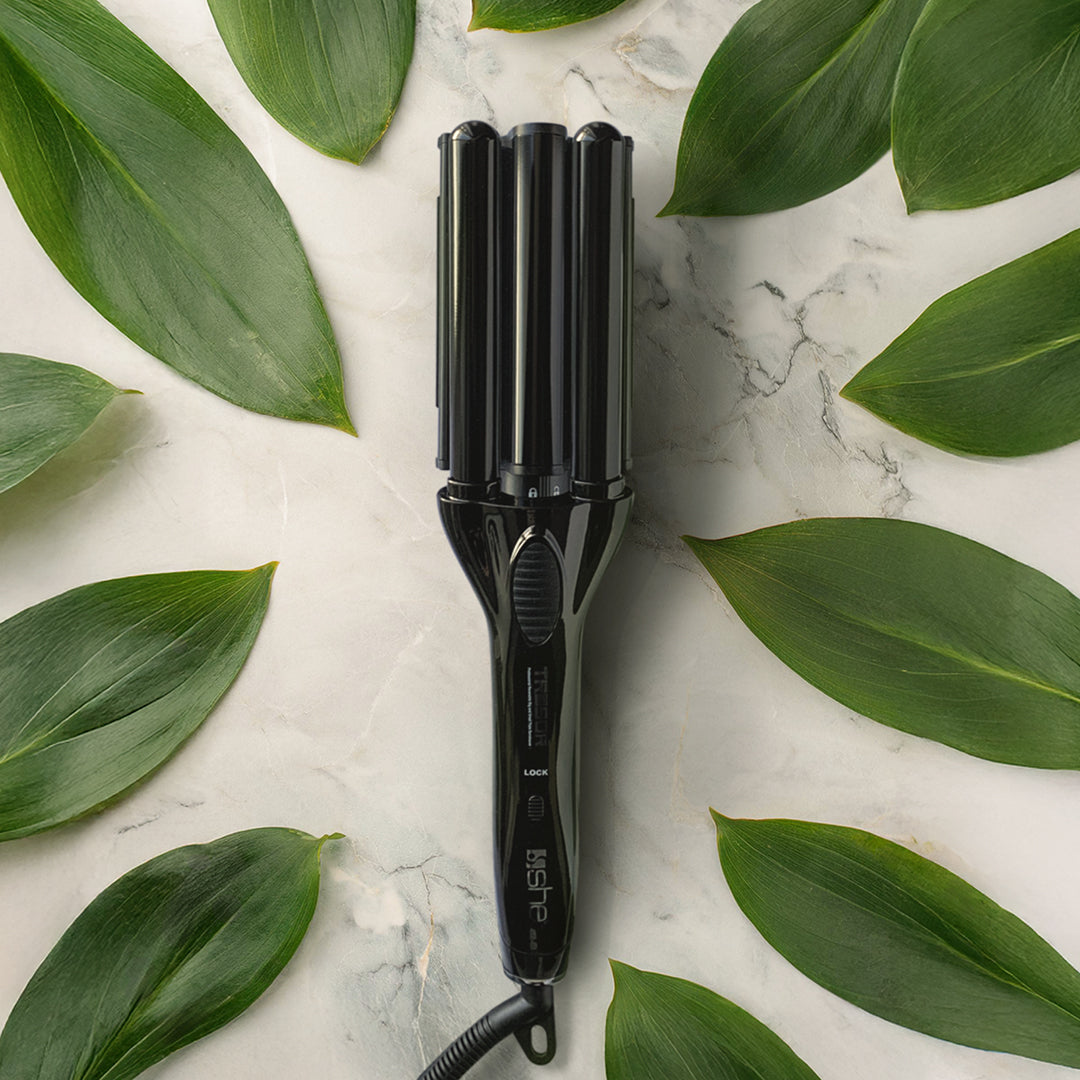 Piastra arricciacapelli nera, che poggia su uno sfondo di marmo bianco con delle venature grigie e foglie verdi