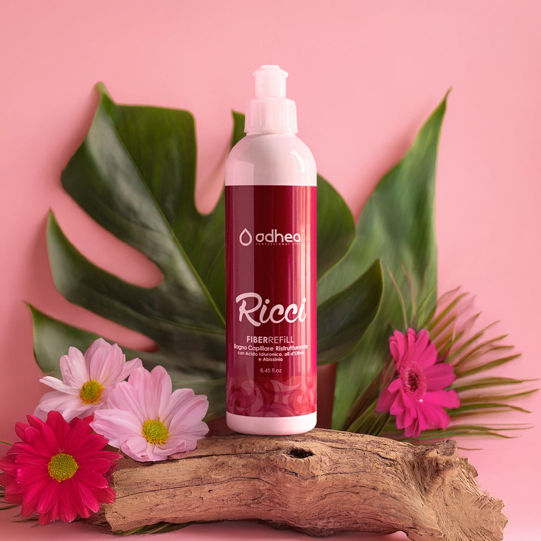 flacone bianco con etichetta rossa, su sfondo rosa e poggia su un pezzo di tronco. Attorno petali rosa e fucsia e si intravede dietro una foglia verde