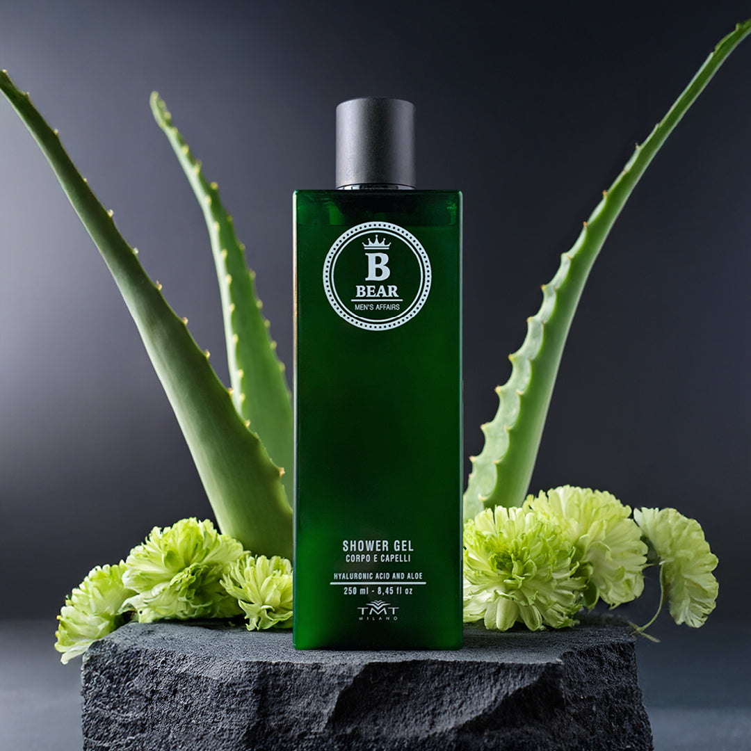 Prodotto Bear dal flacone di vetro con prodotto verde che poggia su un sasso grigio e sfondo grigio. attorno dei fiori verdi e pianta di aloe